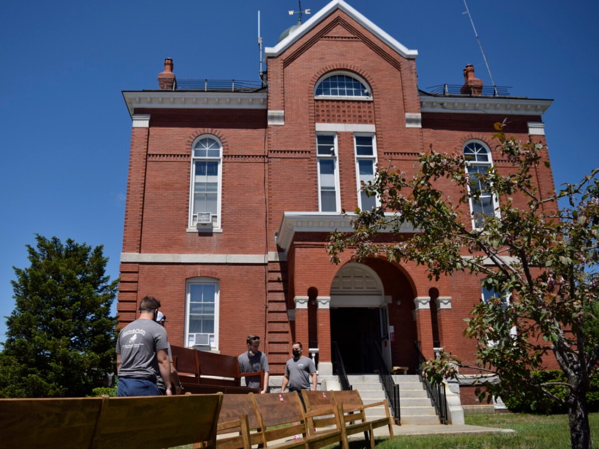 Movers bring in a new cherry-wood stained bench into the Franklin County Superior Court, a red-brick building with white embellishments and big windows. In front of the movers sit benches that are being removed. The old benches are old, worn down, and fading. Behind the courthouse is a bright blue sky without any clouds.